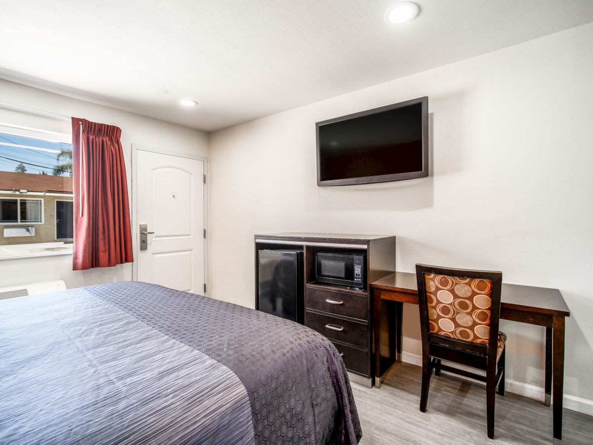 A hotel room with a bed, wall-mounted TV, mini-fridge, microwave, desk with a chair, a window with red curtains, and a door.