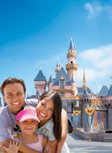 A family of three is smiling and posing in front of a castle in a theme park. The sky is clear and blue in the background, ending the sentence.