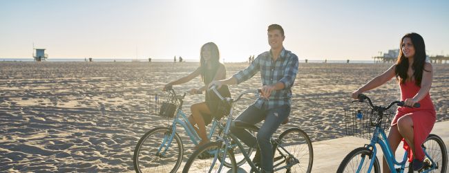 Three people are riding bicycles on a beach boardwalk during sunset or sunrise, with the ocean and sand in the background.