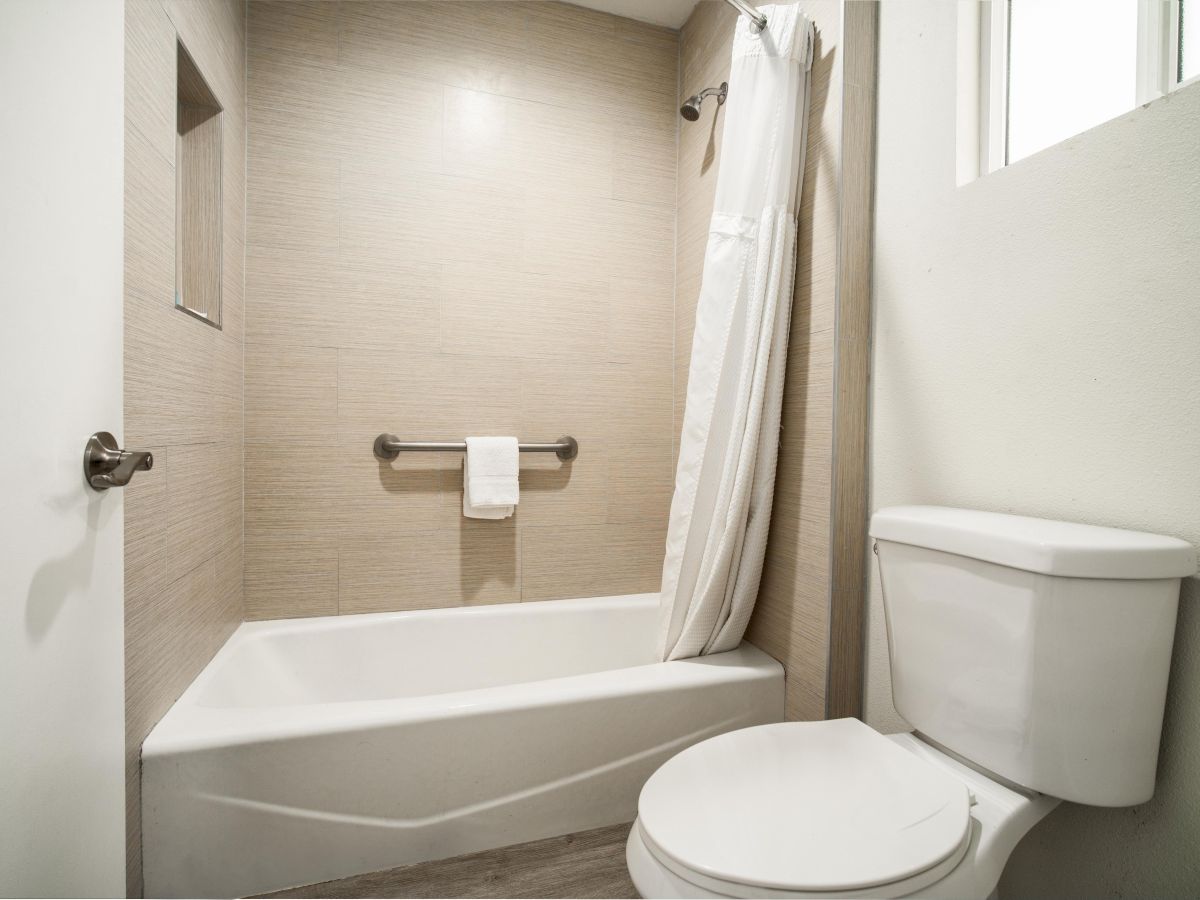 This image shows a clean, modern bathroom with a white bathtub, a curtain, a toilet, a towel rack, and a small window allowing natural light.