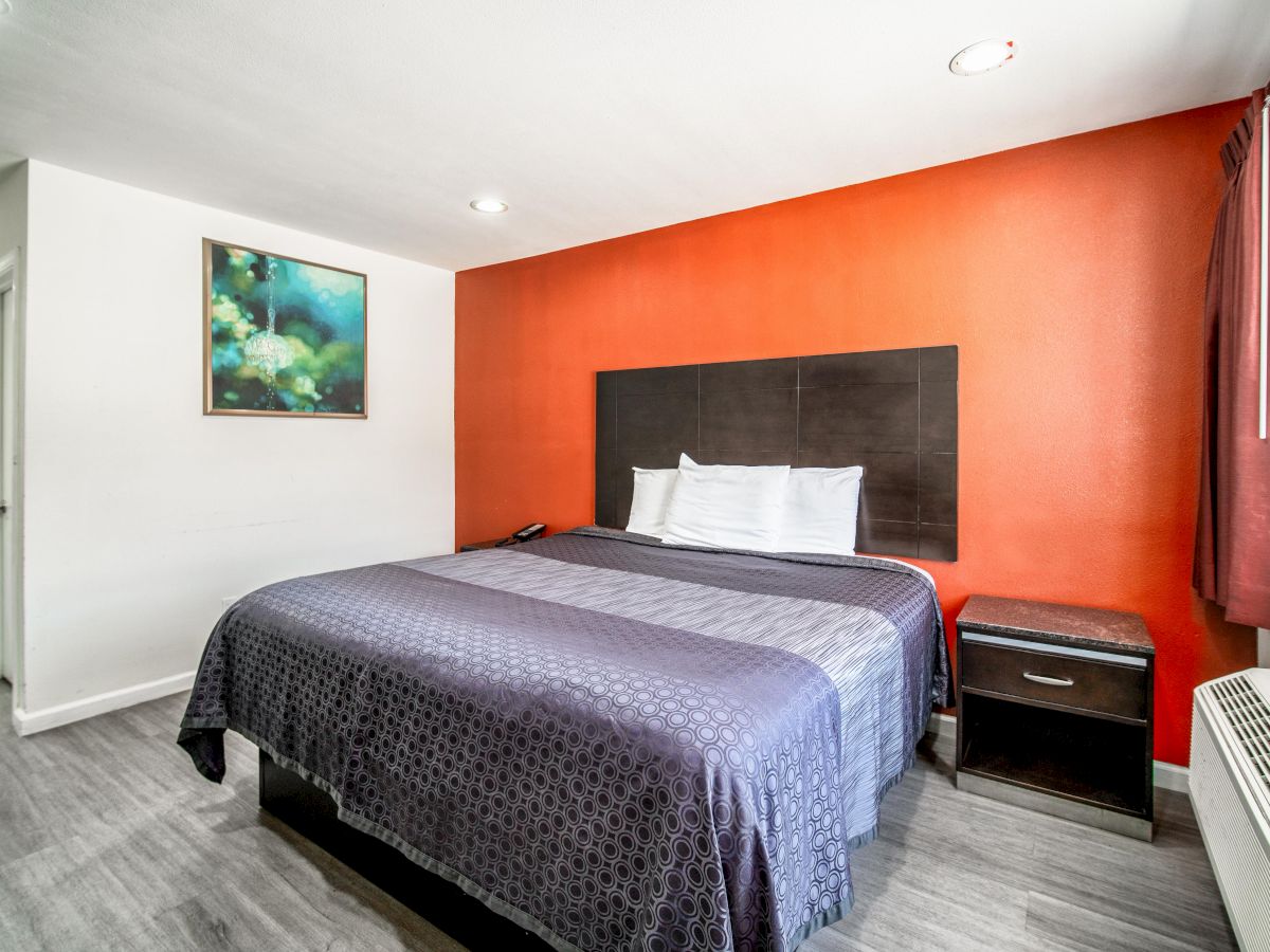 A modern hotel room features a large bed with purple bedding, an orange accent wall, a nightstand, a painting, and a window with curtain.