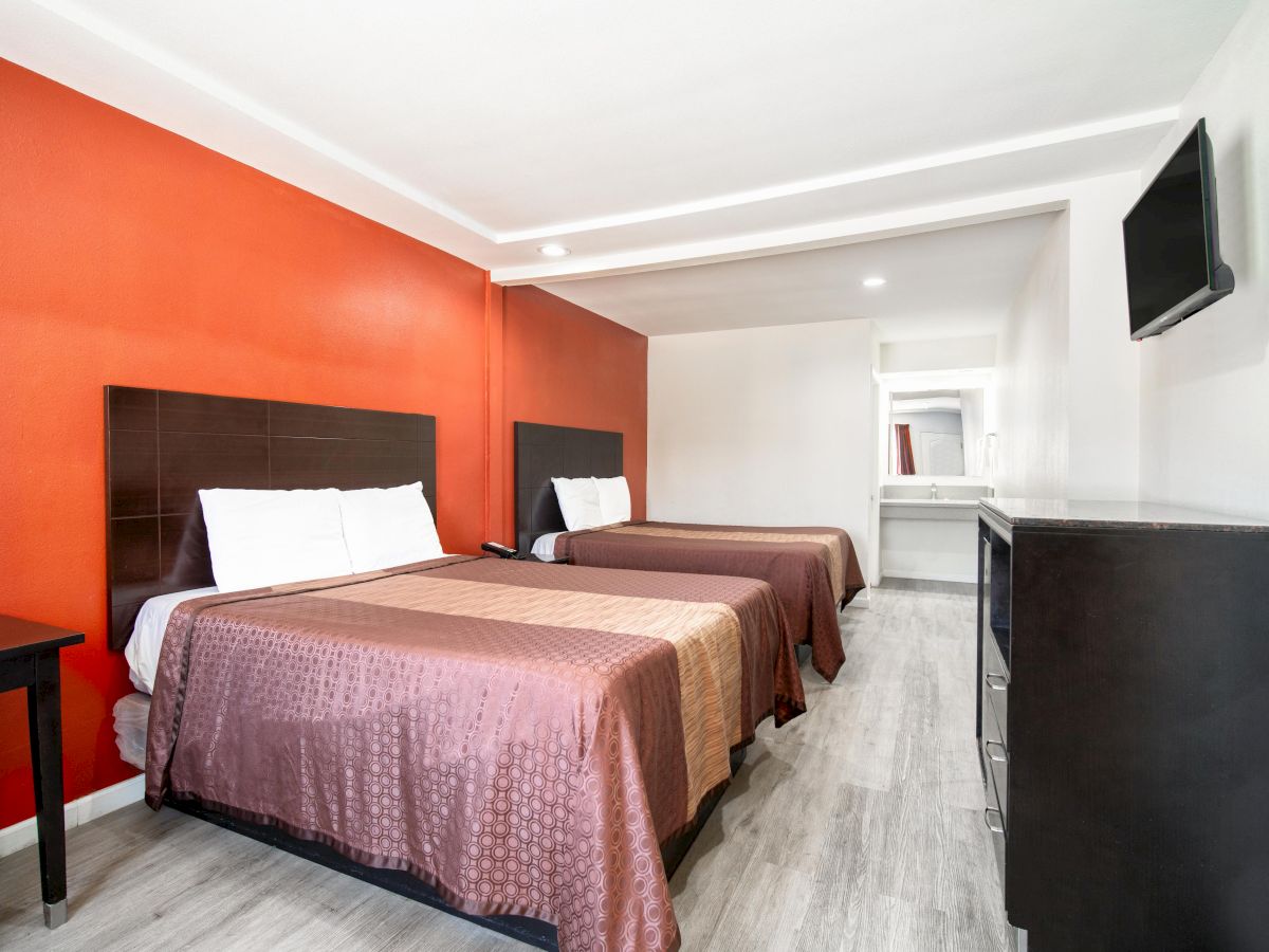 A hotel room with two beds, a wall-mounted TV, a dresser, and a desk. The room has an orange accent wall and a neutral color palette.