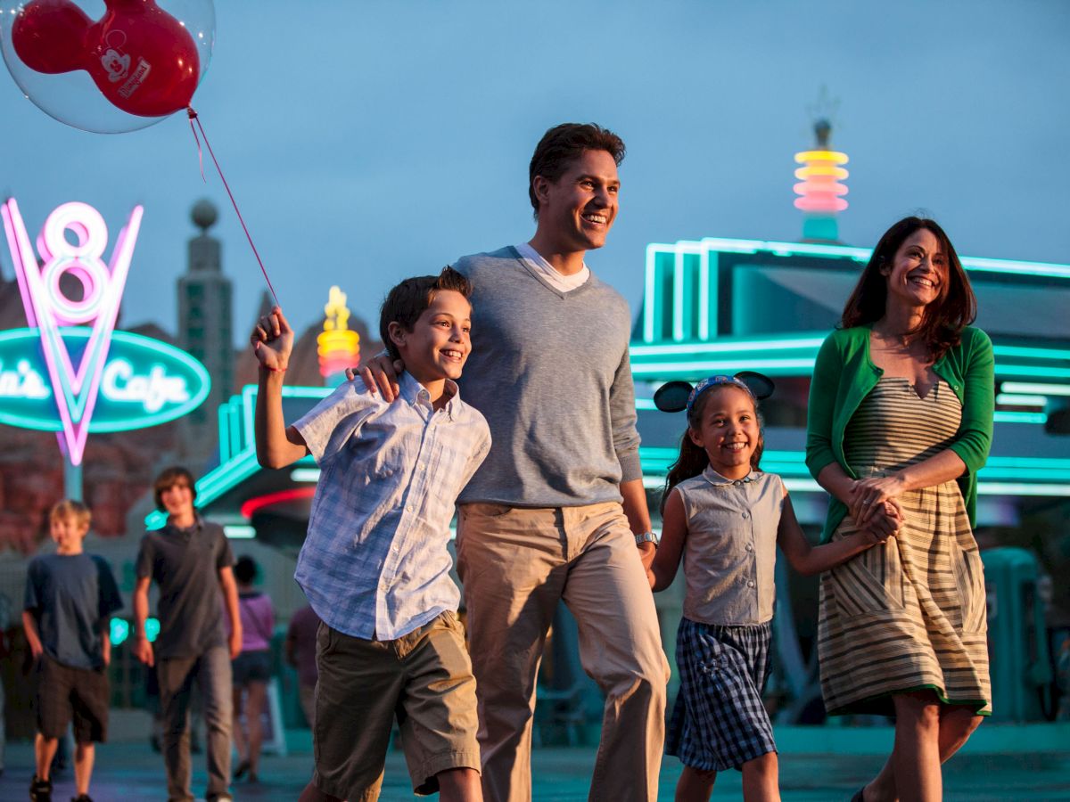 A smiling family of four, with a boy holding red balloons, walks hand in hand in front of a neon-lit diner, surrounded by other people at twilight.