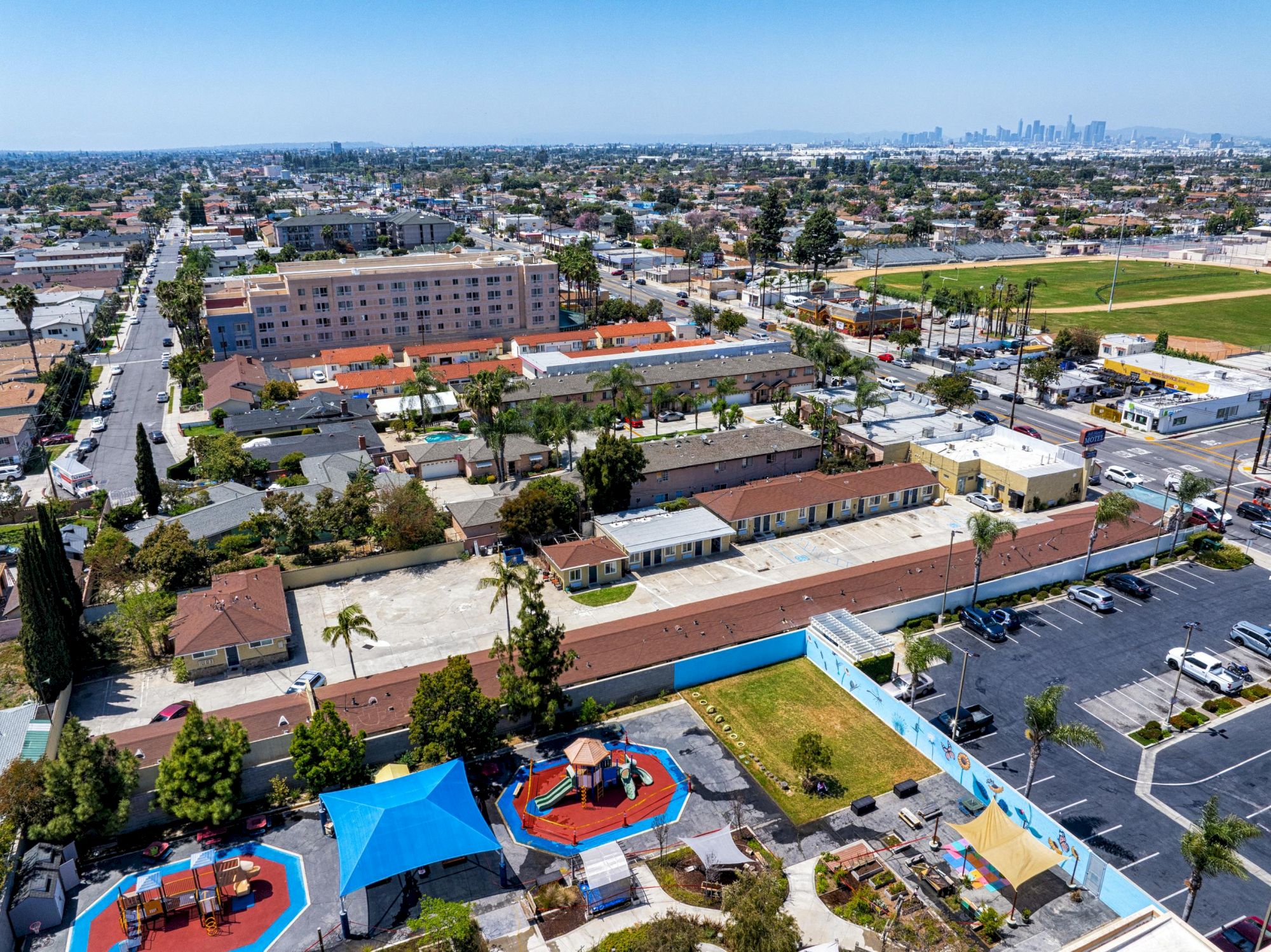 An aerial view of a suburban area featuring a play park, residential houses, buildings, streets, and a distant city skyline in the background.