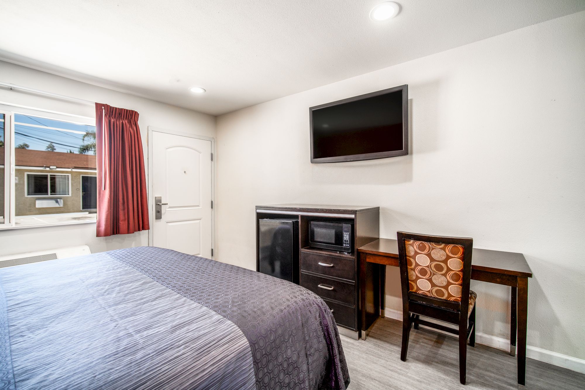 A small hotel room with a bed, TV, mini fridge, microwave, desk, chair, and a window with red curtains, and a white door.