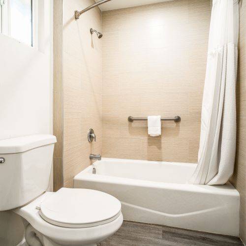 The image shows a clean, modern bathroom with a white toilet, a bathtub with a shower curtain, and a towel rail on a tiled wall.