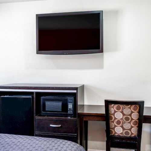 This image shows a hotel room with a flat-screen TV mounted on the wall, a fridge, microwave, desk, and chair with a patterned backrest ending the sentence.