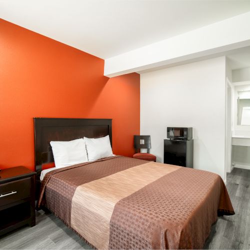 A hotel room with a single bed, two-tone brown bedspread, orange accent wall, nightstand with phone, mini fridge, microwave, and a sink in the back.