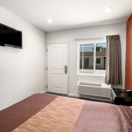 A tidy hotel room with a bed, wall-mounted TV, doorway, window with curtains, air conditioner, and nightstand, featuring a white and red color scheme.