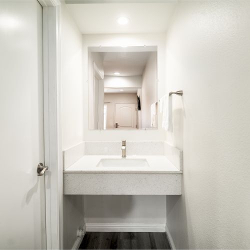 A small, modern bathroom with a minimalist design, featuring a white sink, mirror, towel holder, and a white door.