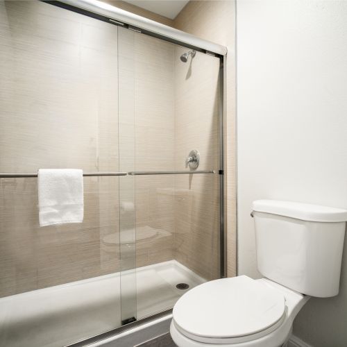 The image shows a bathroom with a glass-enclosed shower, white tiled walls, a white toilet, and a towel hanging on the shower door handle.