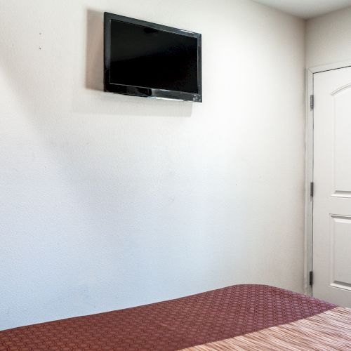 The image shows a minimalistic room with a wall-mounted TV, a portion of a bed with a red bedspread, and a white closed door with a silver handle.