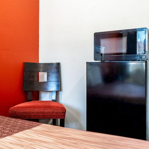 The image shows a small black refrigerator with a microwave on top, a dark chair with a red cushion, and a table against a red and white wall.