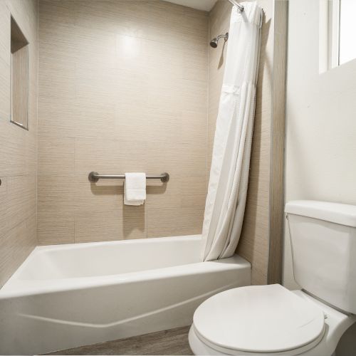 A clean, modern bathroom with a bathtub, white shower curtain, towel rack with a towel, and a white toilet, with neutral wall and floor tiles.