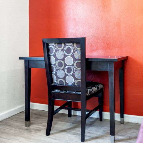 A simple workspace with a black desk and chair, backed by an orange wall, next to an air conditioning unit and partially visible bed.