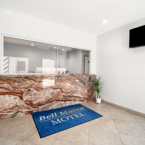 A motel reception area with a marble desk, a potted plant, a wall-mounted TV, and a blue mat that says 