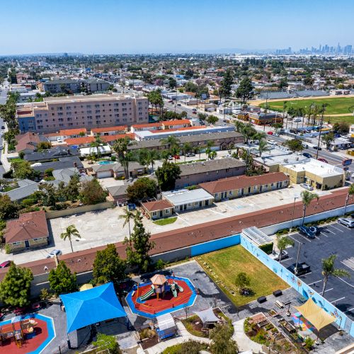 This image shows an aerial view of a suburban area with residential buildings, a playground, a parking lot, and a distant city skyline.