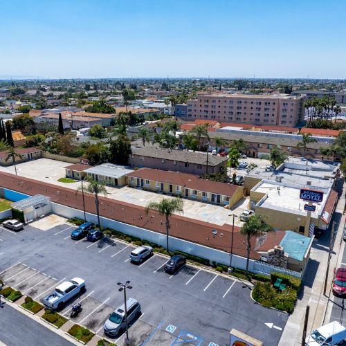 Aerial view of an urban area with buildings, parking lot, and a busy road on the right. Trees and cars are visible.