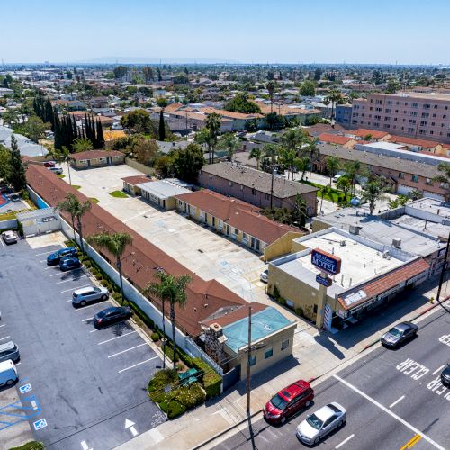 Aerial view of an urban area featuring a parking lot, streets with cars, buildings, and a motel with a sign. Trees and various structures surround it.