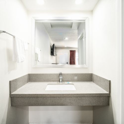 The image shows a modern bathroom with a sink, a large mirror, and towels hanging on a rack to the left. The background shows part of a room.